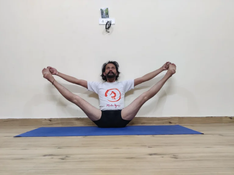 A man demonstrating Upavistha Konasana B, or Wide-Angle Seated Forward Bend variation, in yoga with hands clasped behind his back.